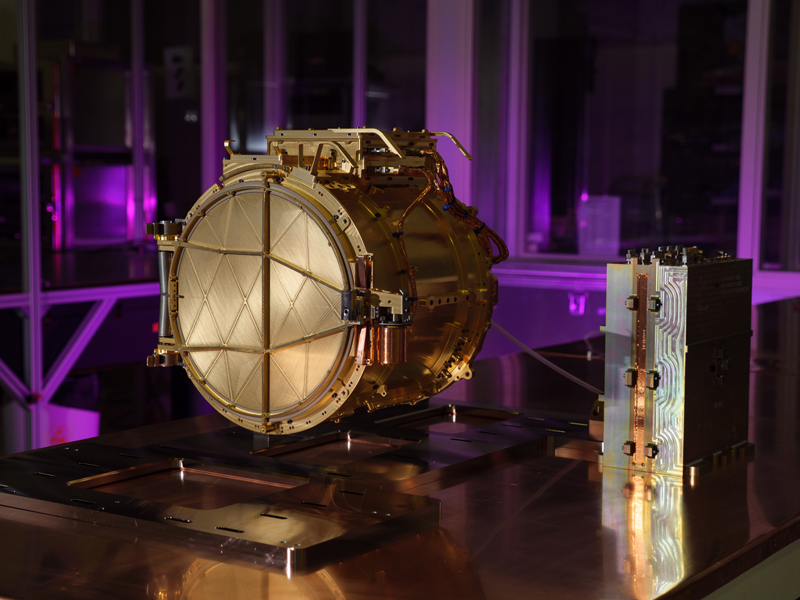 The gold colored SUDA sensor head is shown resting on a table in a clean room, seen in the center left of this image. The cover to the sensor head is closed, and to the right a small, silver electronics box is visible. Combined, the pieces make up the entire SUDA instrument. The background space in the clean room is lit up purple. 