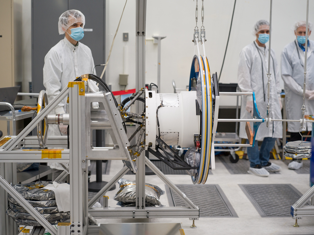 Three workers in white lab coats, cleanroom bouffant caps, and blue face masks watch as Europa Clipper’s magnetometer boom is tested. Part of the equipment holding the boom can be seen in front of the workers. 