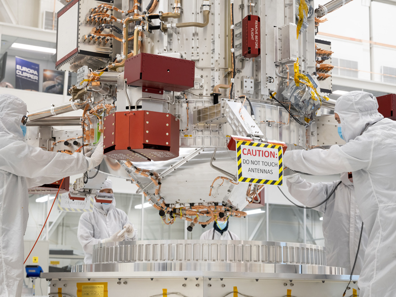 The base of the main body of the Europa Clipper spacecraft is seen in the middle of the image. The main body is large, silver, and shaped like a cylinder, with wiring and electronics attached. Four people are visible surrounding the main body of the spacecraft, wearing full body coveralls and masks. The people are gently guiding the spacecraft into its stand where it will continue to be worked on in the Spacecraft Assembly Facility.   