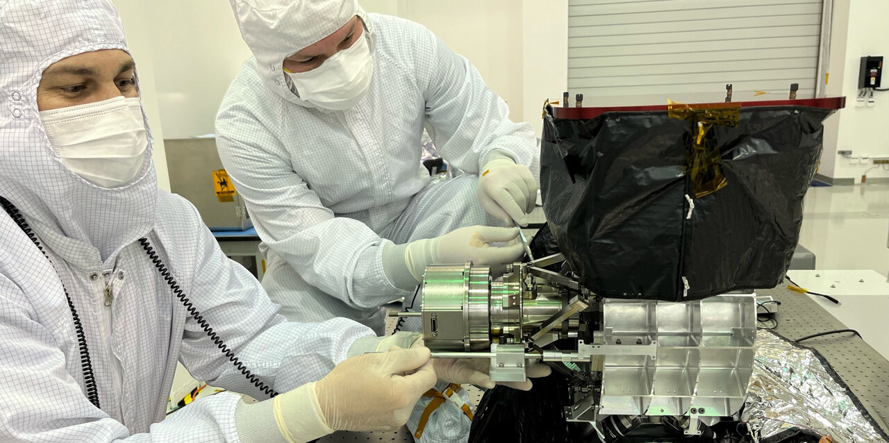 Two engineering flight technicians, one with white and one with light-brown skin, wearing full protective gear at a table in a clean room, installing the flight scanner into the MISE instrument.