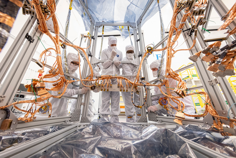 people wearing clean room attire work on the spacecraft's electrical system
